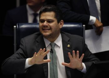Homem de terno escuro e gravata verde senta em cadeira azul, gesticulando com as mãos abertas à frente do corpo. Ao fundo, outras pessoas vestem roupas formais, uma segura papéis brancos.
O presidente do Senado, Davi Alcolumbre (União Brasil-AP)