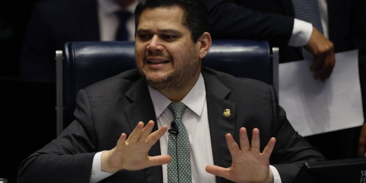 Homem de terno escuro e gravata verde senta em cadeira azul, gesticulando com as mãos abertas à frente do corpo. Ao fundo, outras pessoas vestem roupas formais, uma segura papéis brancos.
O presidente do Senado, Davi Alcolumbre (União Brasil-AP)