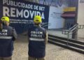 Dois agentes da Ordem Pública do Rio, com coletes azuis e bonés amarelos, observam área onde propaganda externa foi removida. Letreiro grande indica remoção de publicidade irregular em espaço público com piso de pedra portuguesa e escadaria ao fundo.
Agentes da Prefeitura do Rio de Janeiro removem anúncio de bet em espaço público