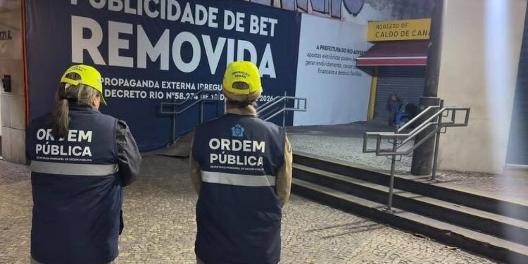 Dois agentes da Ordem Pública do Rio, com coletes azuis e bonés amarelos, observam área onde propaganda externa foi removida. Letreiro grande indica remoção de publicidade irregular em espaço público com piso de pedra portuguesa e escadaria ao fundo.
Agentes da Prefeitura do Rio de Janeiro removem anúncio de bet em espaço público